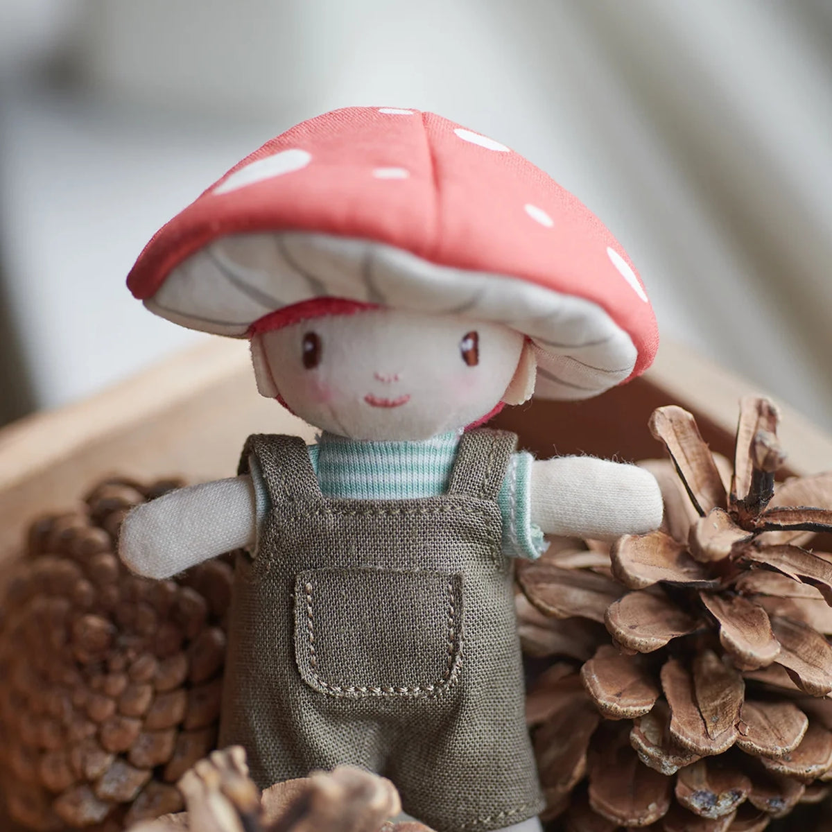 Small plush doll with a red toadstool hat sitting among natural pinecones.