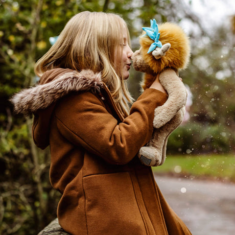 A young blonde girl in a winter coat holds up a plush creature, kissing its nose outdoors in a park.