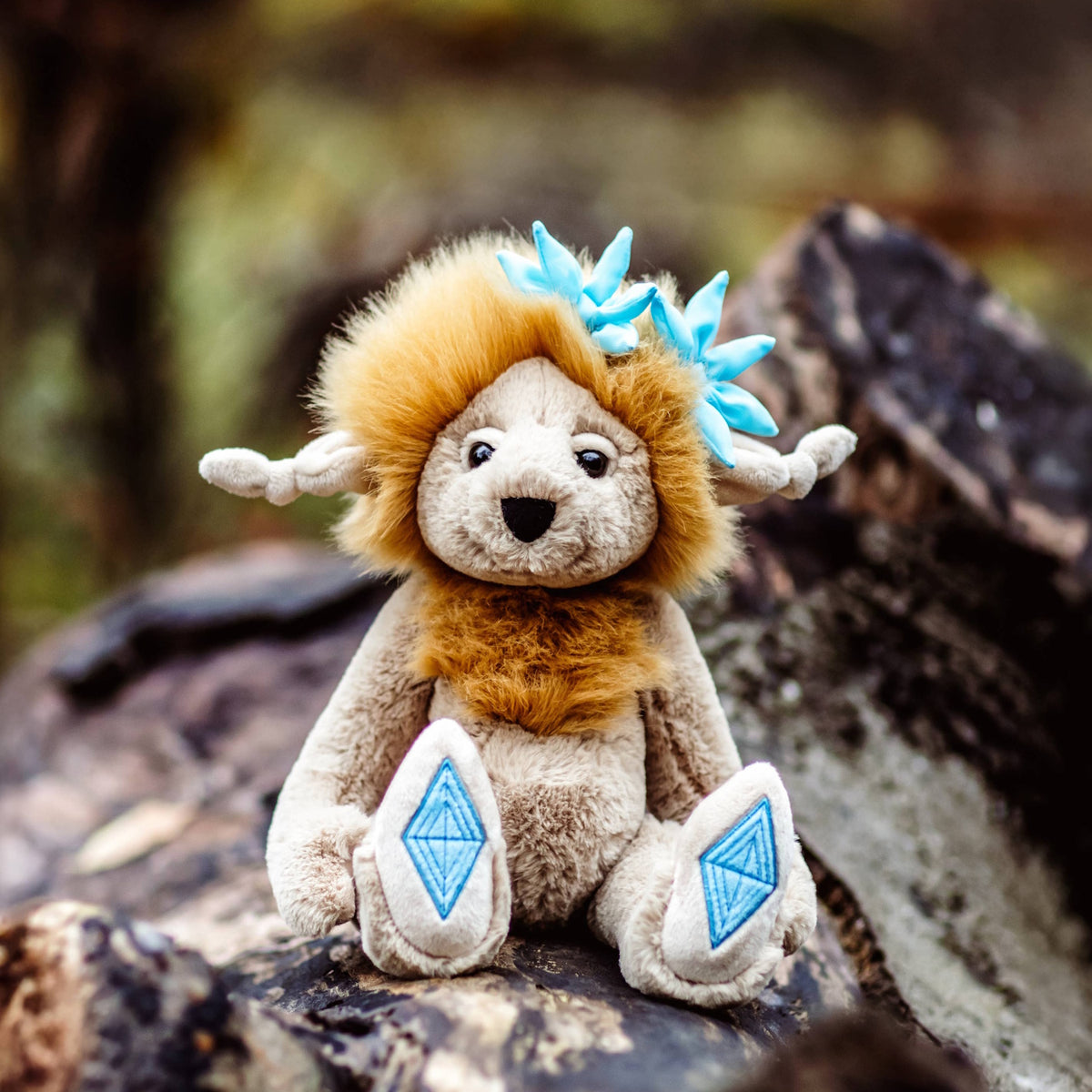 Plush creature with a golden mane and blue flowers sitting outdoors on a wooden log with blurred foliage behind.