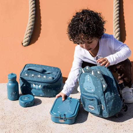 Young child playing with a matching blue triceratops backpack, lunchboxes, and water bottle outside.