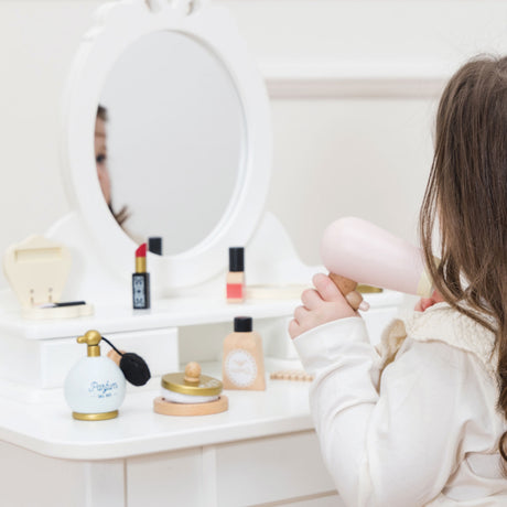 Child using a toy wooden hair dryer at a white vanity table set with play cosmetics.