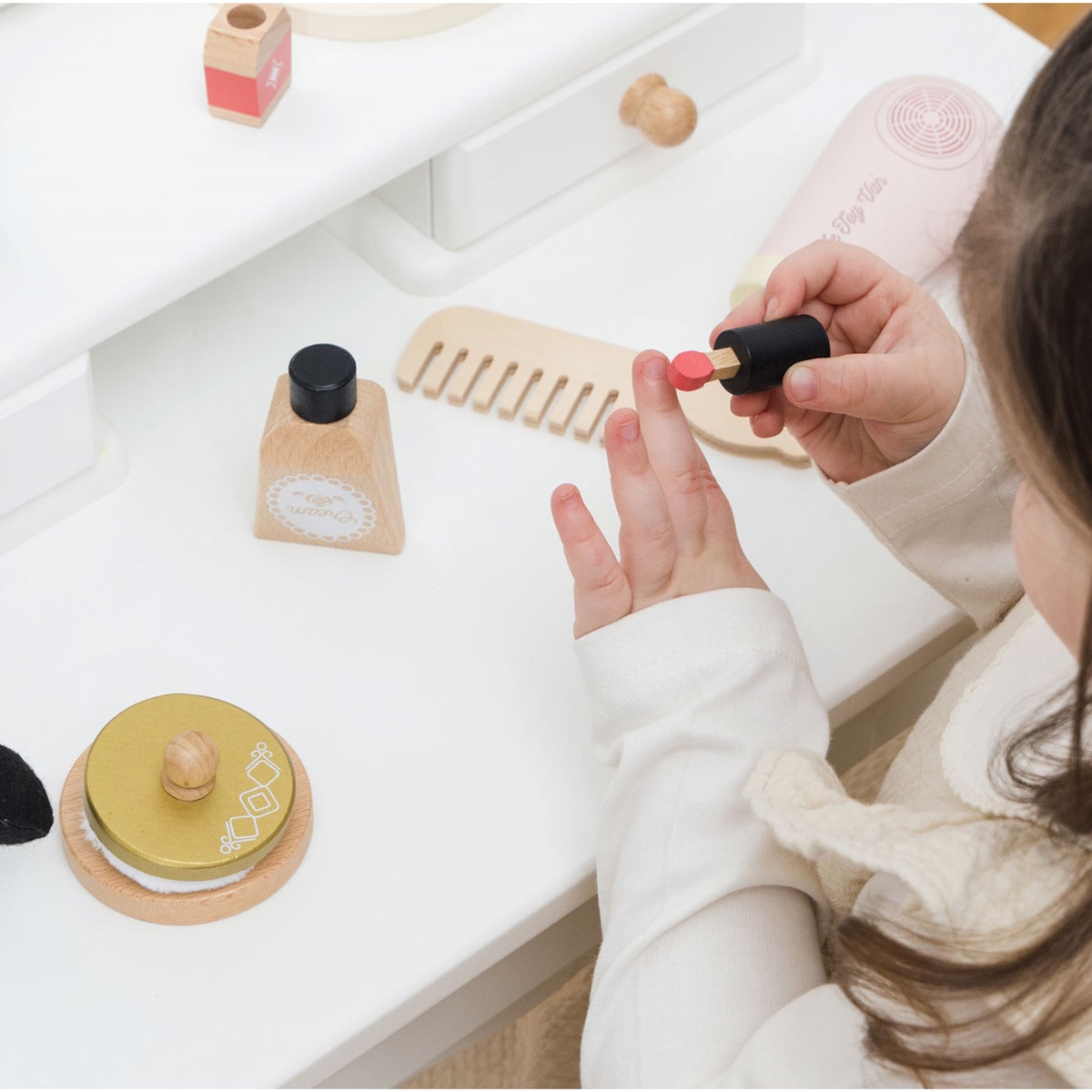 Child playing with a wooden toy lipstick at a white vanity table with beauty accessories.