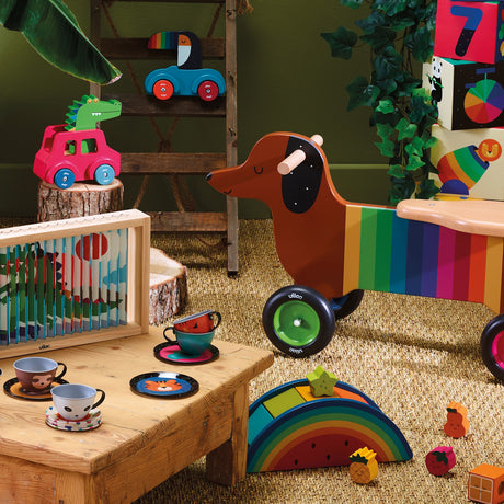 Colorful wooden children's toys including a rainbow dachshund rider, animal tea set, and blocks on a natural rug.