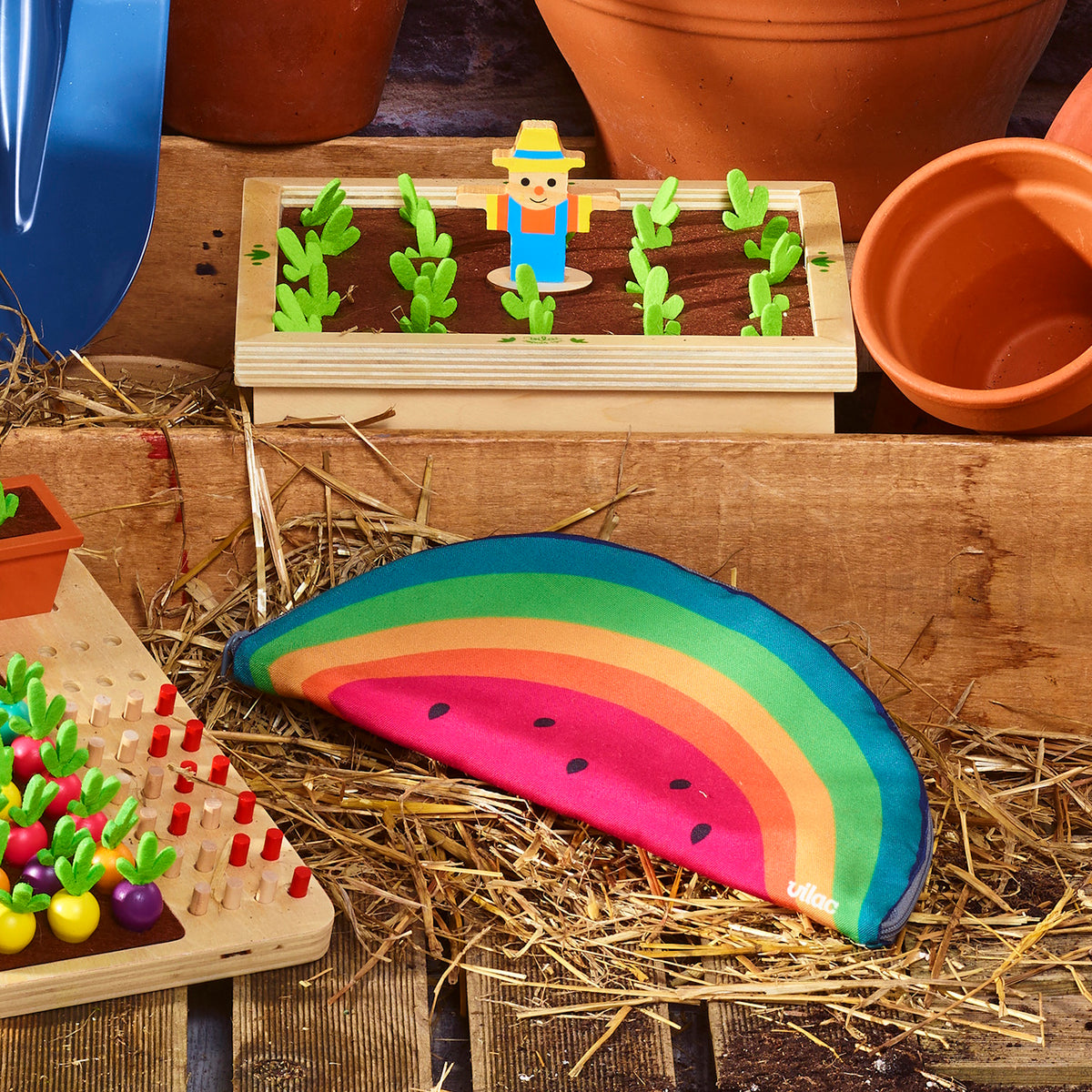 Vilac rainbow watermelon pouch resting on straw next to wooden garden toys and flower pots.