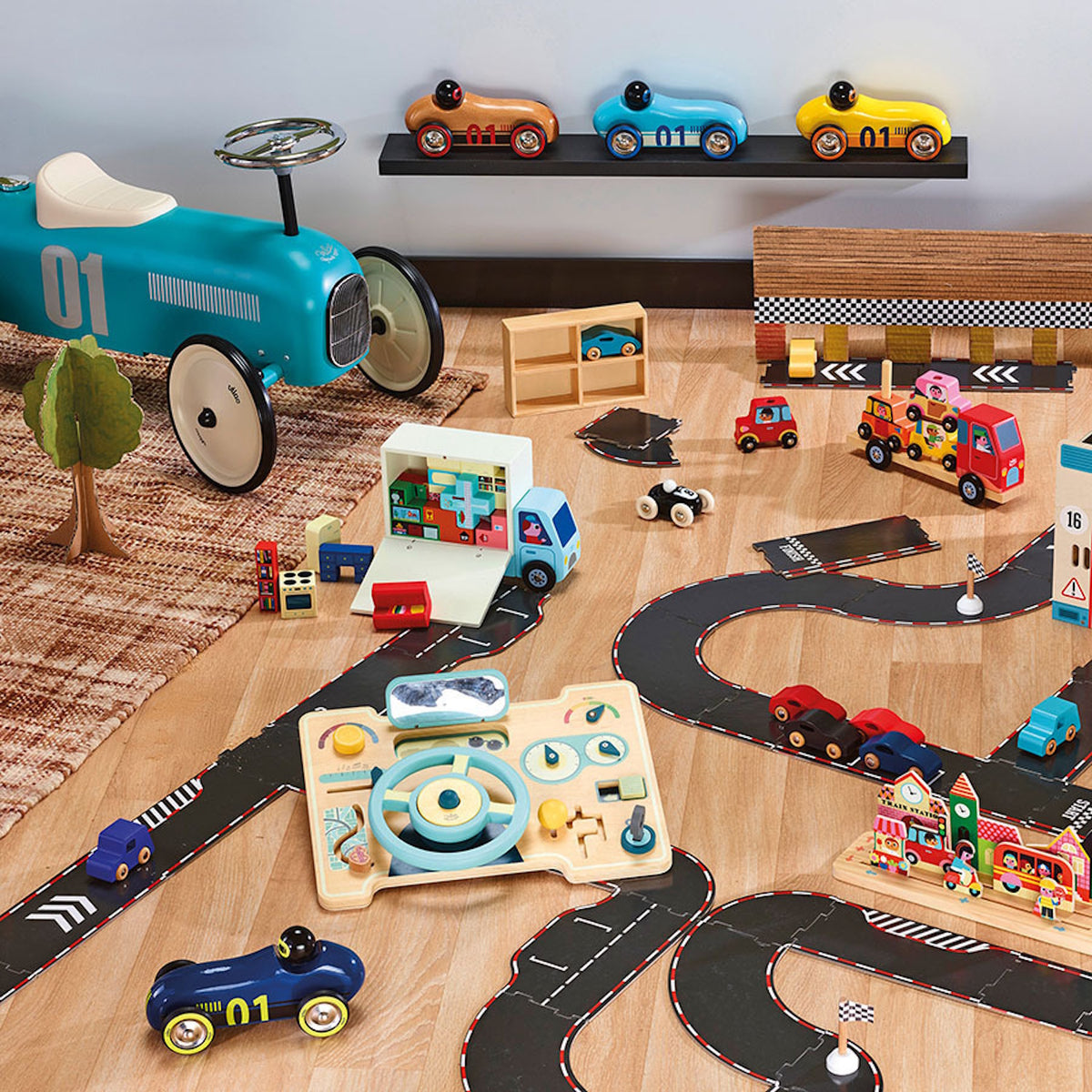 Wooden toy cars, blue ride-on car, and a toy moving truck on a play road track on a playroom floor.