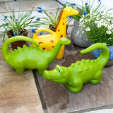 Green dinosaur, yellow giraffe, and lime crocodile animal-shaped watering cans on a stone patio with potted garden flowers.