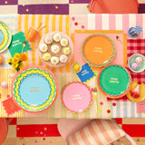 Overhead view of a colorful, patterned birthday table setting with vibrant paper plates and cupcakes.