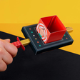 A hand holds a colorful, rectangular, paper-made device resembling a sword-swallowing magic trick, set against a black and yellow background.