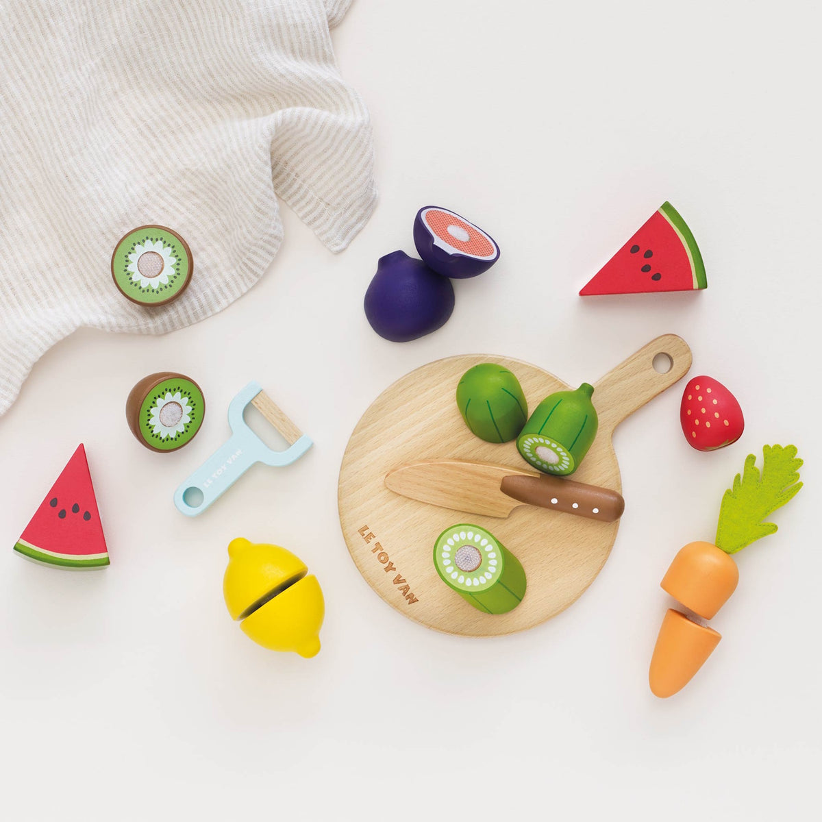 Wooden toy play food pieces arranged around a chopping board with a toy knife and cloth.
