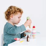 A young child with curly blonde hair playing with a wooden toy ice cream cone and stand set at a table.
