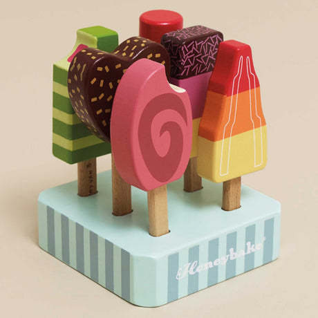 Six colorful wooden toy ice lollies/popsicles set upright in a striped light-blue base on a beige background.