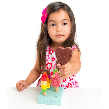 A young girl wearing a floral dress holding up a chocolate heart-shaped wooden play popsicle, with the set's stand in front of her.
