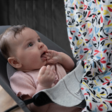 A baby wearing a pink outfit lies in a gray bouncer, gazing intently at a colorful patterned blanket. The scene conveys curiosity and wonder.