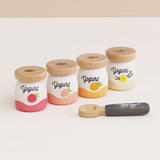Four wooden play yogurt pots arranged diagonally with wooden lids and a toy spoon in the foreground.